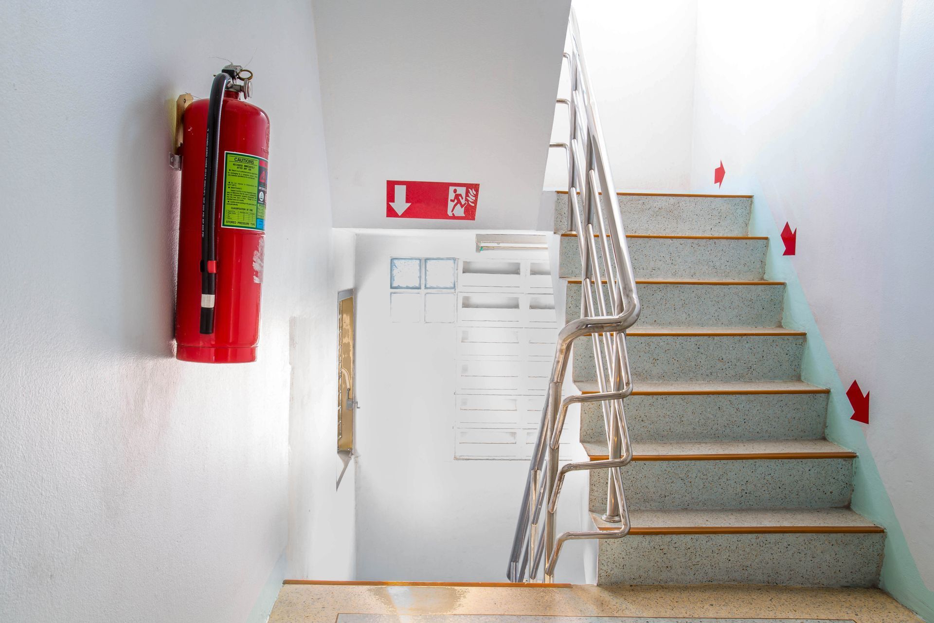 Red Fire Extinguisher on White Wall Next to Staircase With Emergency Exit Signs — Industry Fire Services in Cape York, QLD