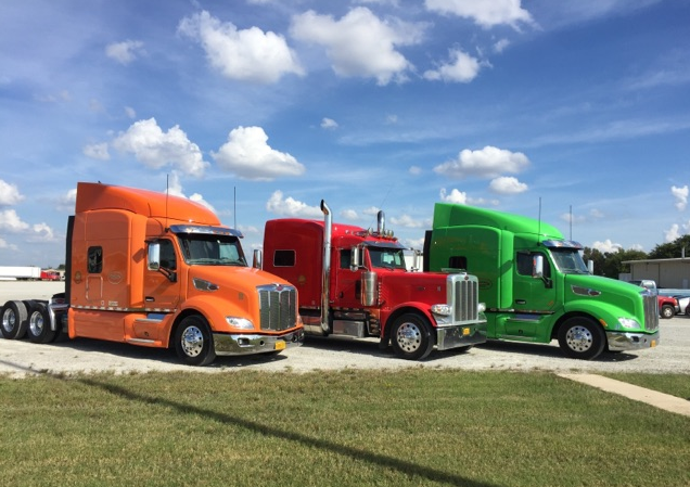 Three colorful semi trucks—orange, red, and green—parked side by side under a blue sky.