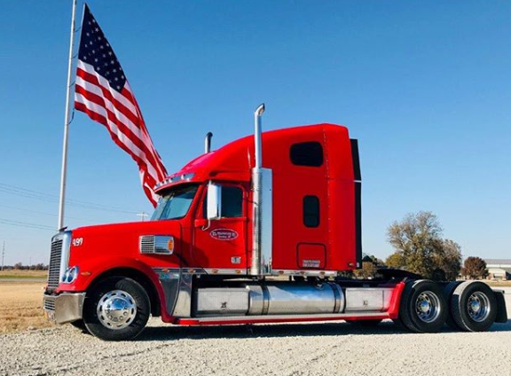 Red semi truck with American flags parked on a roadside under a clear blue sky