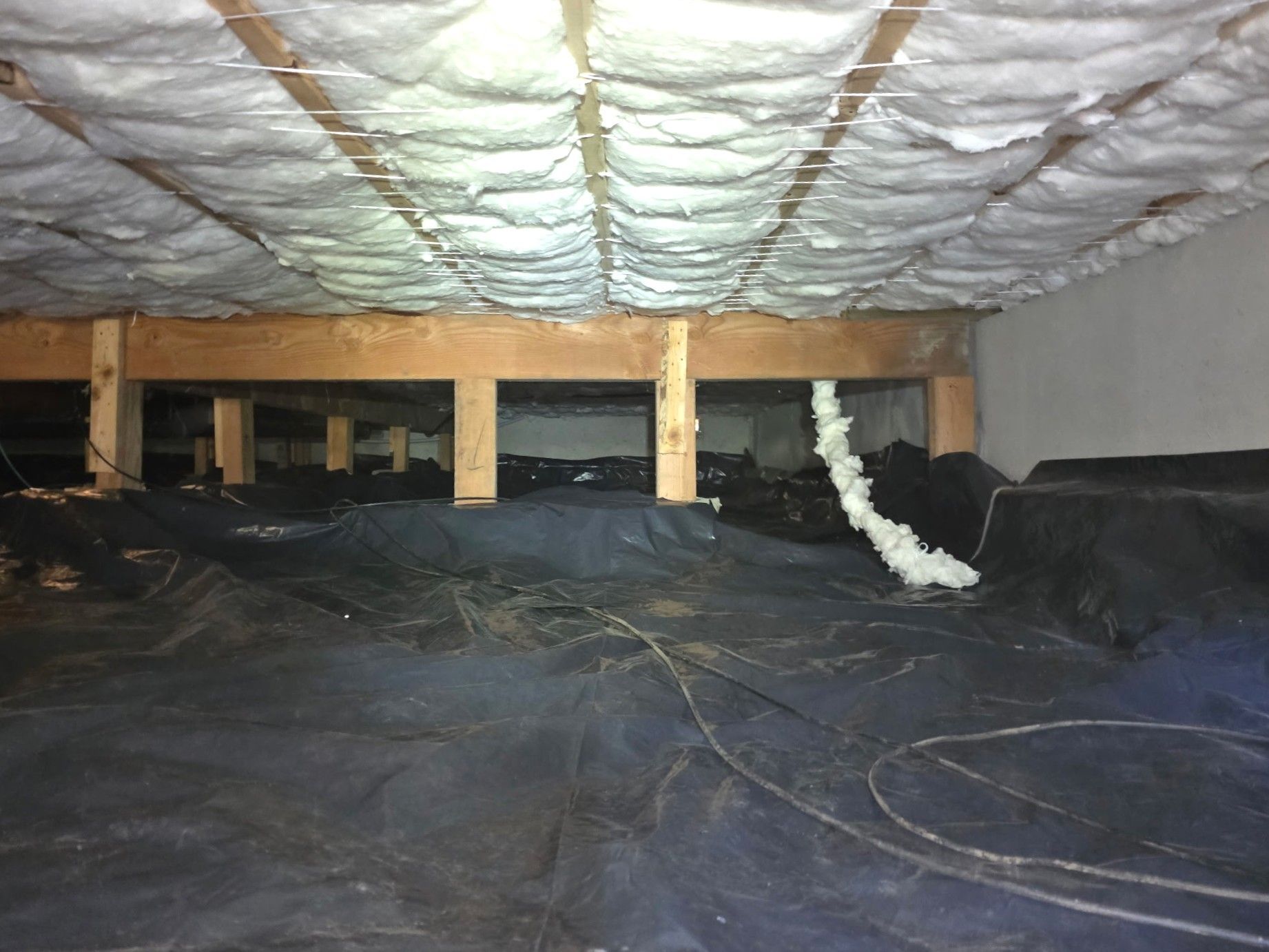 A crawl space with a dark vapor barrier on the ground, wooden floor joists, and white insulation installed overhead.