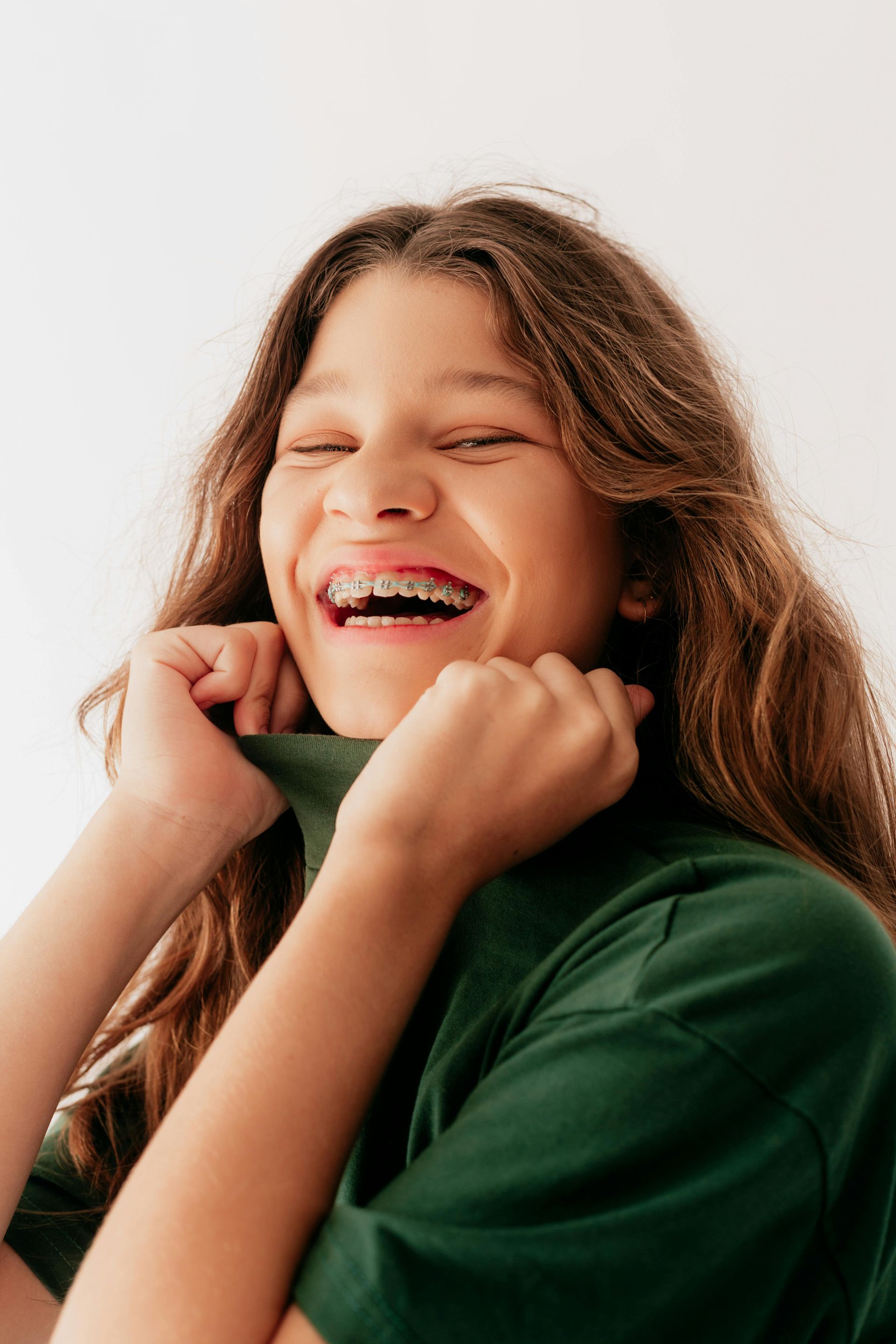 Smiling teen with braces in a green shirt, holding the collar up, with eyes closed.