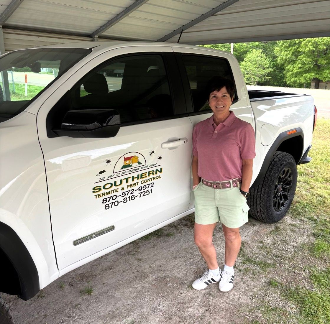 Woman standing beside white pickup truck under a covered area, wearing pink shirt and shorts.