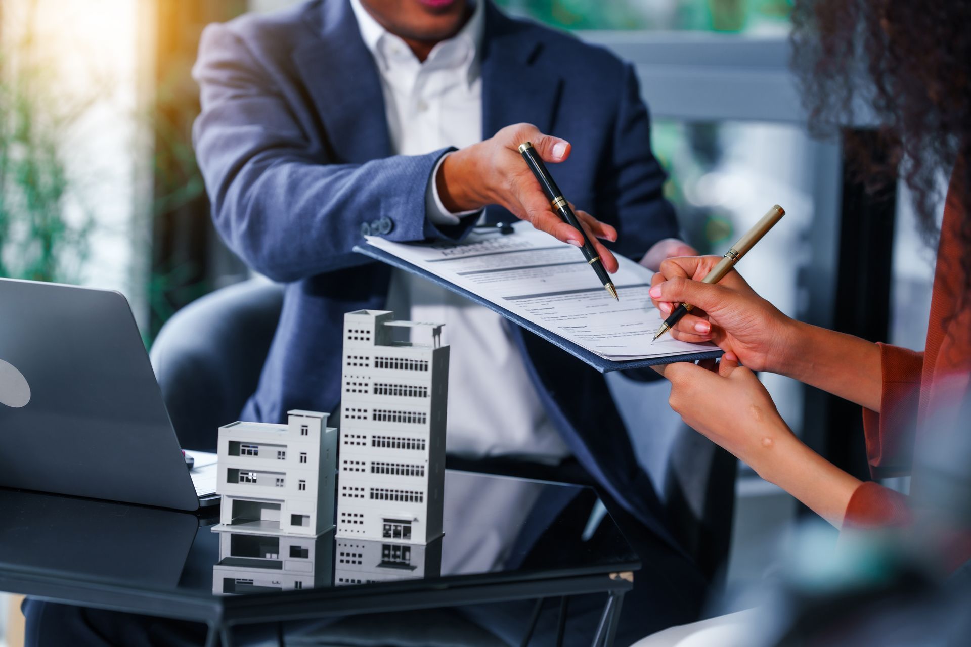 Broker points to a contract as an investor signs it beside commercial building models.