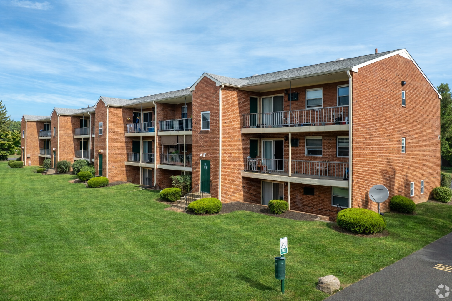 A three-story red brick apartment building with recessed balconies and a green lawn under a blue, cloudy sky.