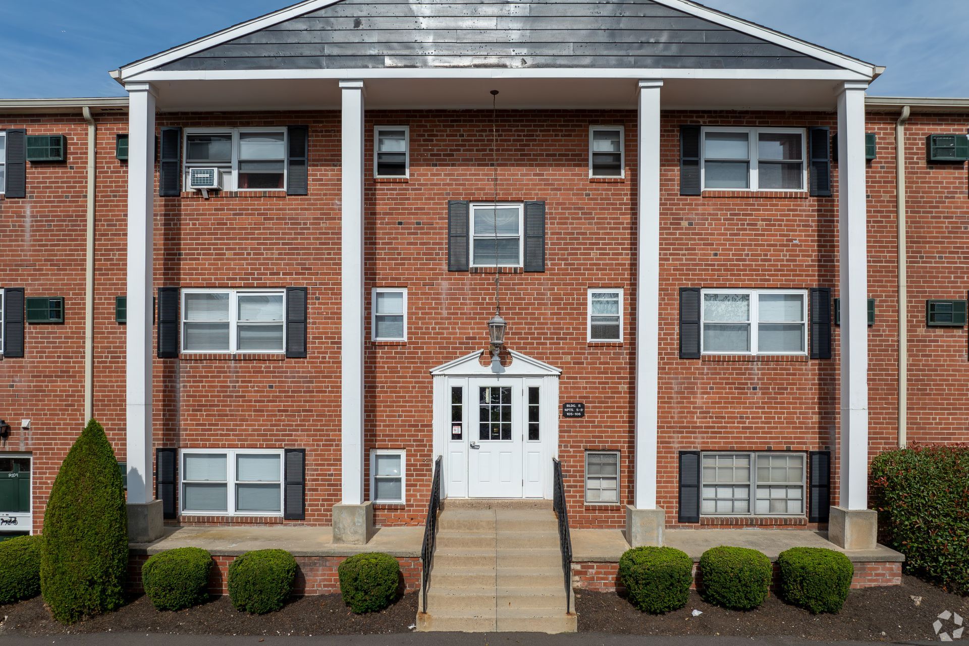 A three-story red brick apartment building with a white front entrance, central pillars, and neatly trimmed landscaping.