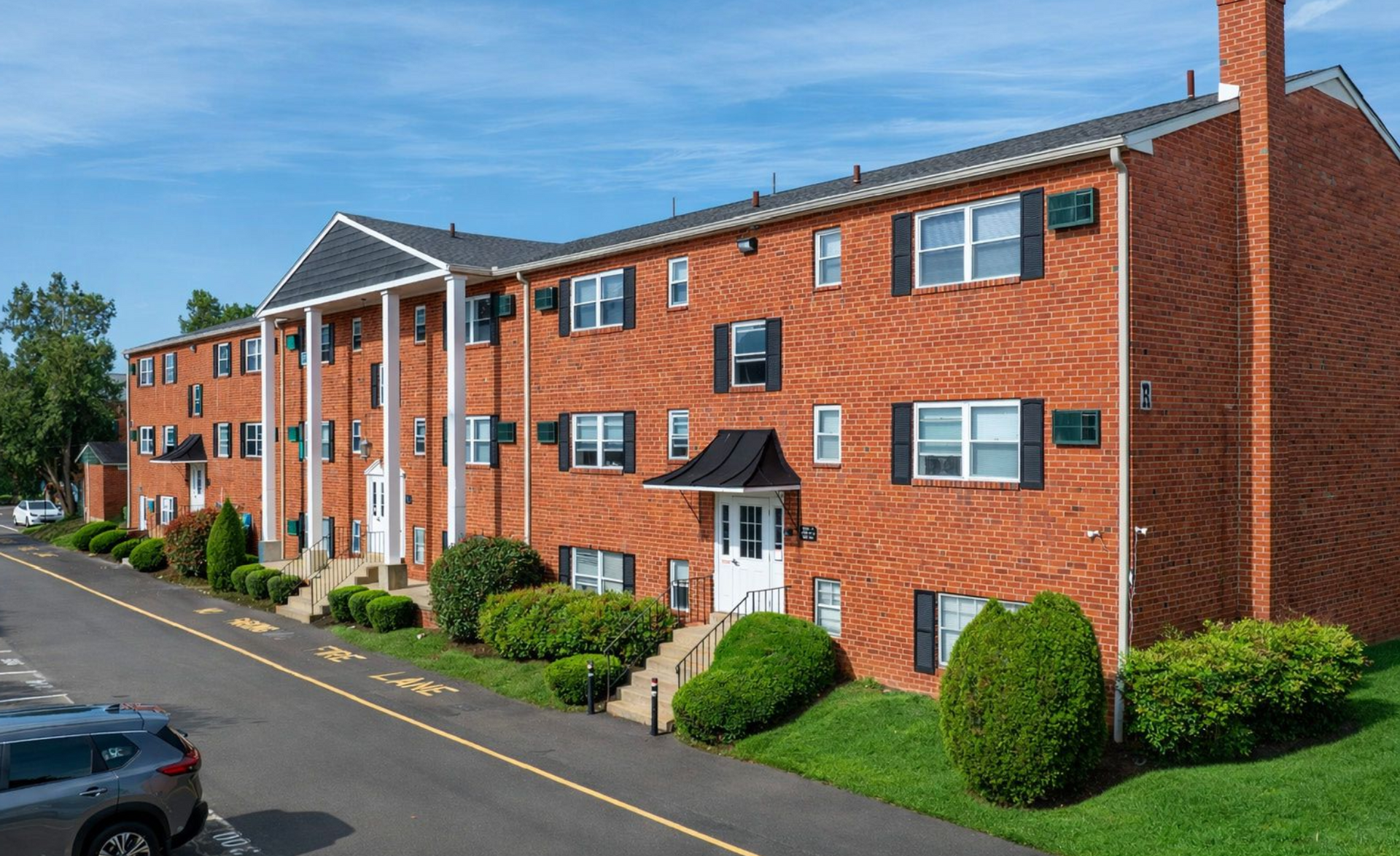 Three-story red brick apartment building with white columns at the entrance, a chimney, and a small parking lot.
