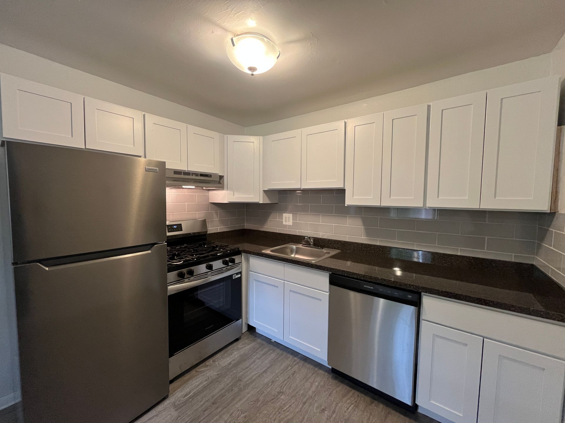 A bright kitchen with white cabinets, stainless steel appliances, dark countertops, and light wood-look flooring.