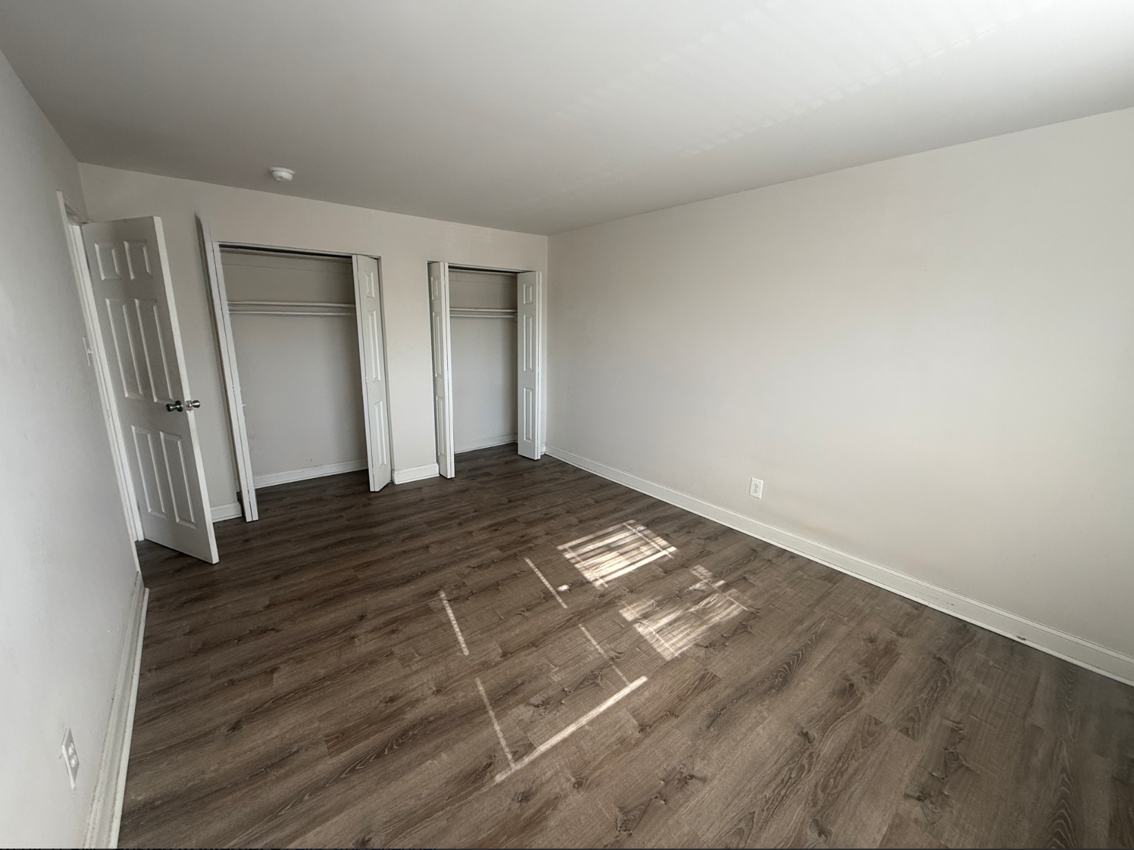 A vacant room with light grey walls, dark wood-look floors, and two open closets with white doors.