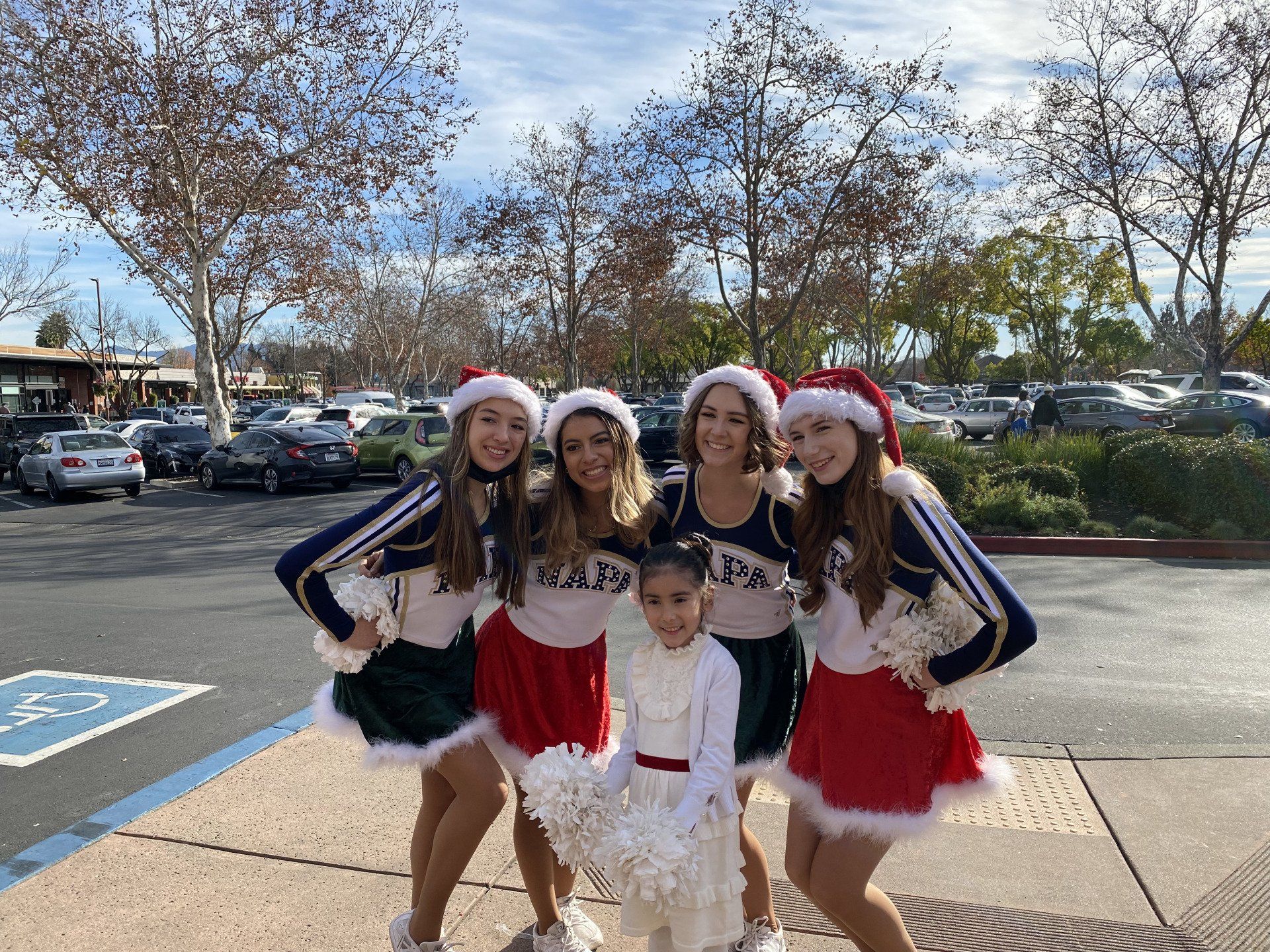 A group of cheerleaders are posing for a picture with a little girl in a parking lot.