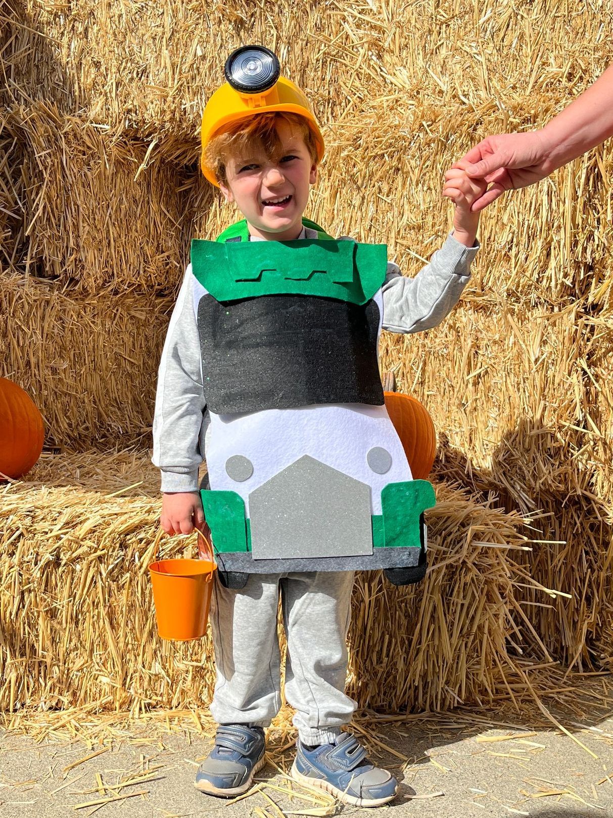 A young boy in a construction worker costume is standing in front of hay bales.