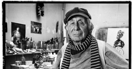 Zwart-witportret van een persoon met een gestreepte sjaal en muts in het atelier van een kunstenaar.
