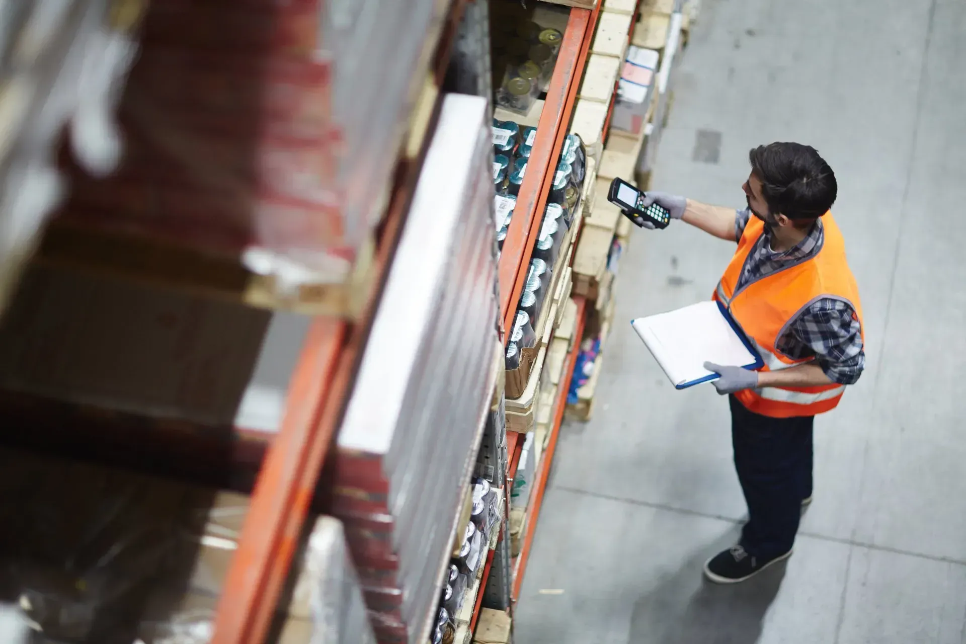 Worker in orange vest using a handheld scanner while checking shelves in a warehouse aisle