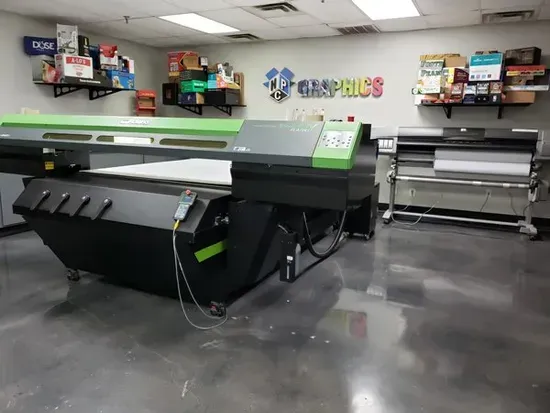 Large-format printer and cutter machines in a print shop workspace, with supplies on wall shelves.