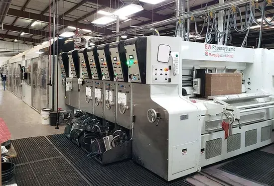 Large industrial printing press in a factory, with multiple metal units and control panels under bright overhead lights
