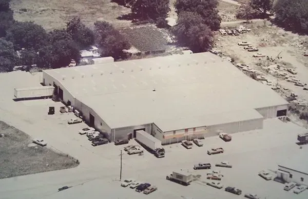 Aerial view of a large beige warehouse with parked vehicles and trees around it.