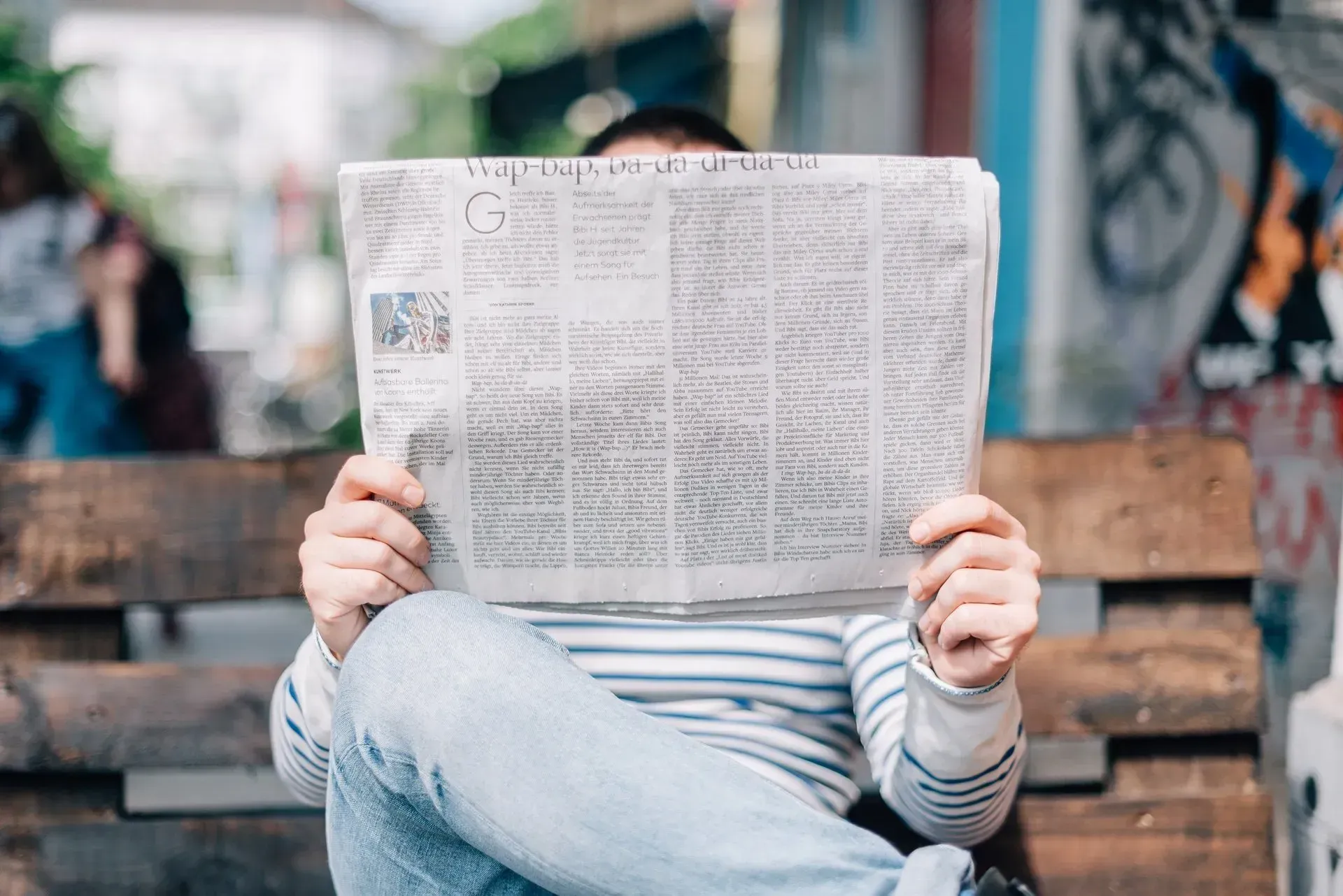 Person sitting on a bench reading a newspaper, face hidden behind it, outdoors.