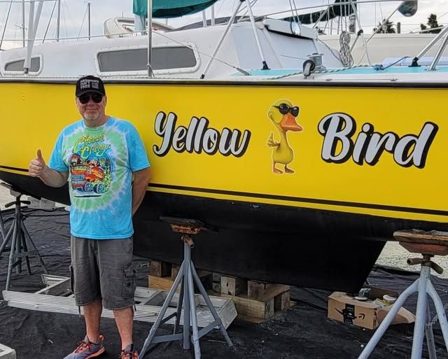 A man is standing in front of a yellow boat that says yellow bird
