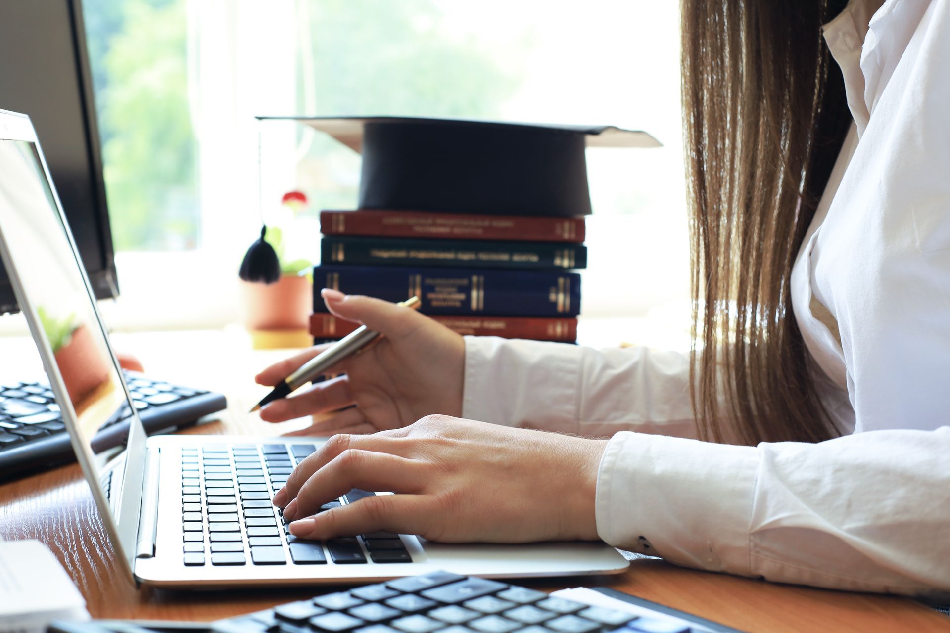 Person typing on a laptop at a desk with a graduation cap and books in the background