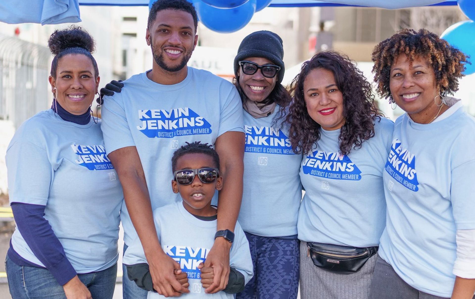 Six people in blue and white T-shirts posing outdoors, smiling with a banner in the background.