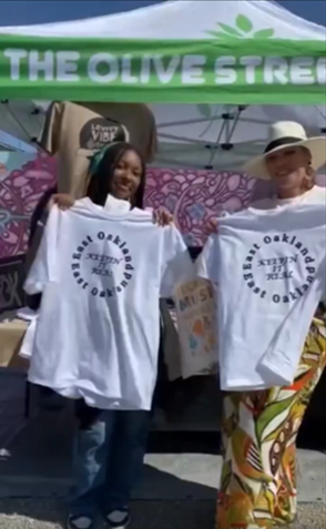 Two people holding white T-shirts at an outdoor market booth under a green “THE OLIVE STR…” canopy