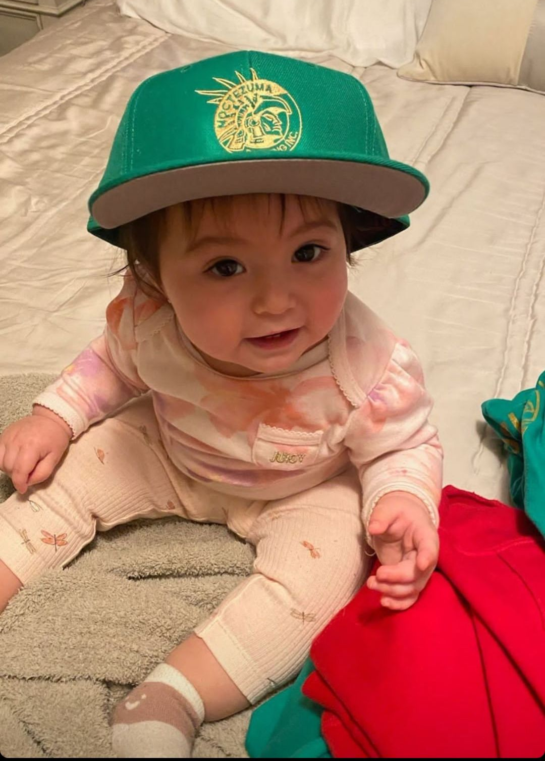 Baby sitting on a bed wearing a green cap and pink outfit, looking at the camera.
