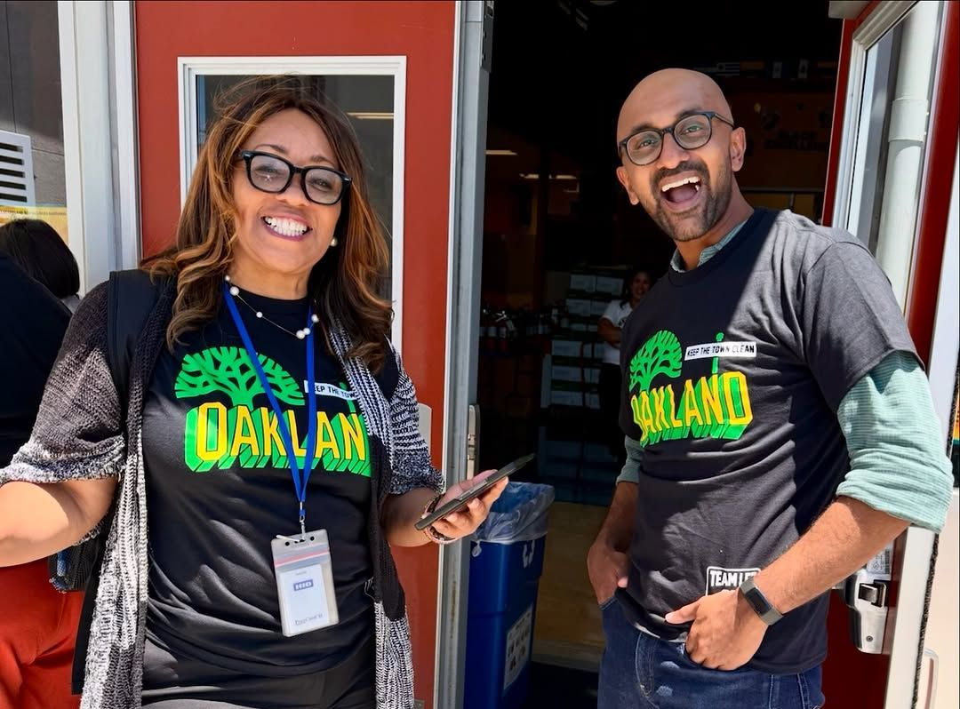 Two smiling people in Oakland shirts standing in a doorway, one holding a phone