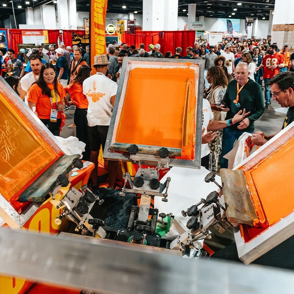 People gathered around a screen-printing machine at a busy indoor convention.