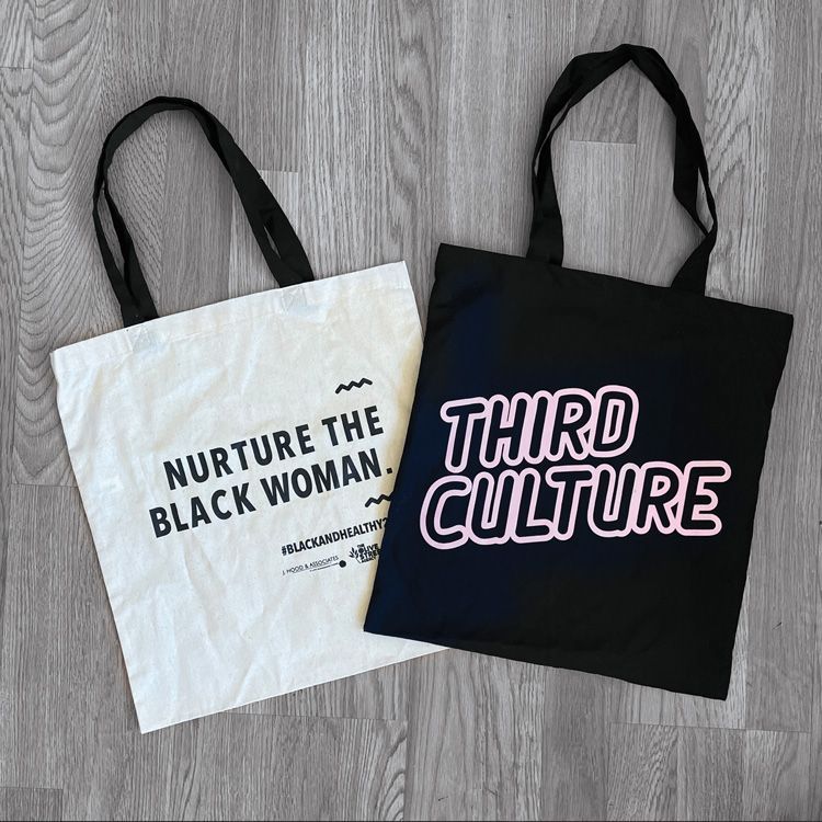 Two tote bags on a wood floor: white bag says “Nurture the Black Woman,” black bag says “Third Culture”