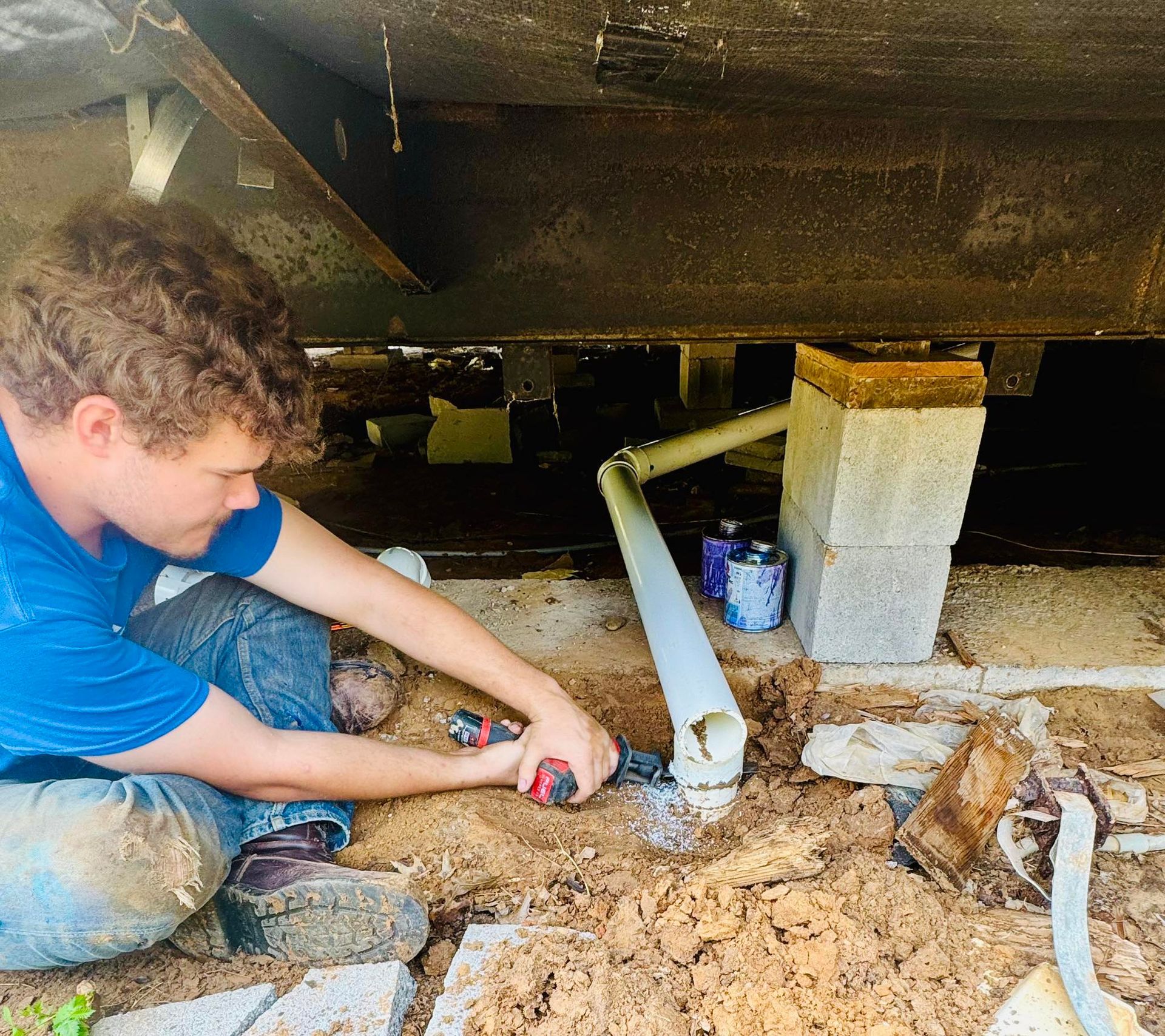 A person works on PVC pipes under a structure, using a drill. Outdoor setting, ground visible.