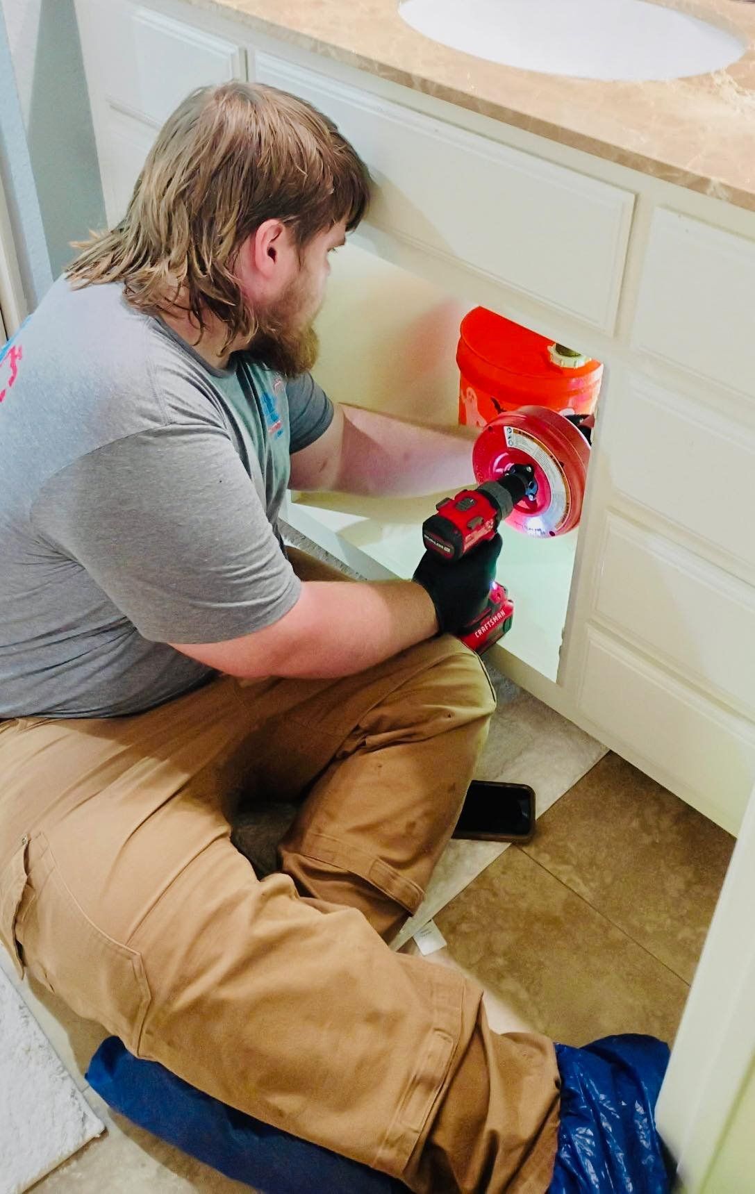 Man using a drill on a white cabinet under a sink. He is wearing work clothes. Orange bucket nearby.
