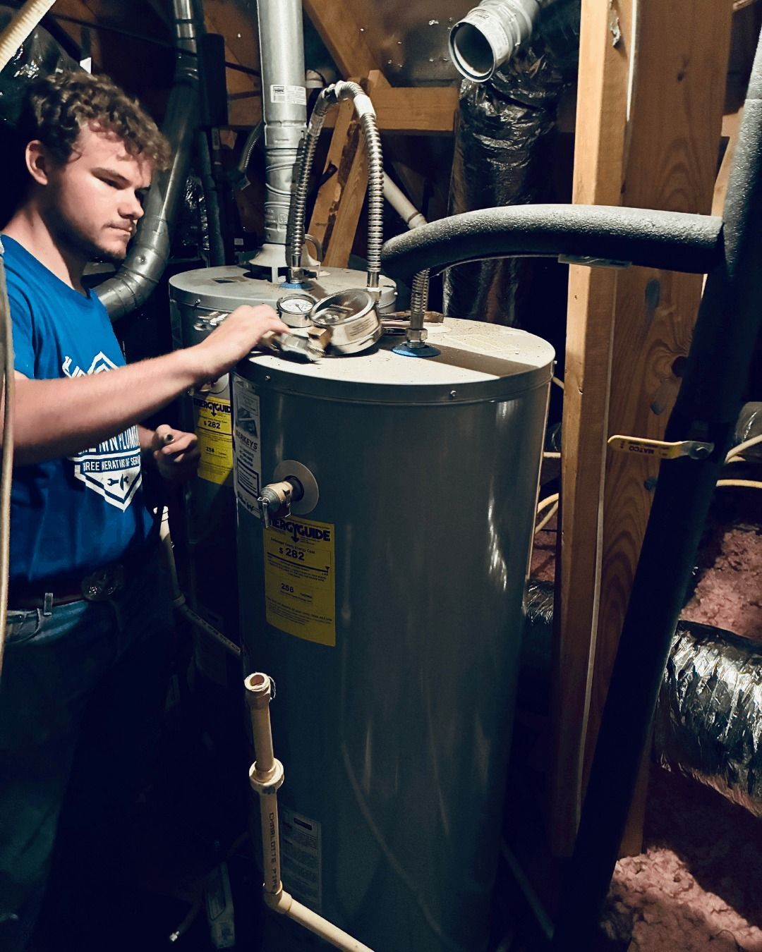Person working on a water heater in an attic. He is wearing a blue shirt and working on the tank.