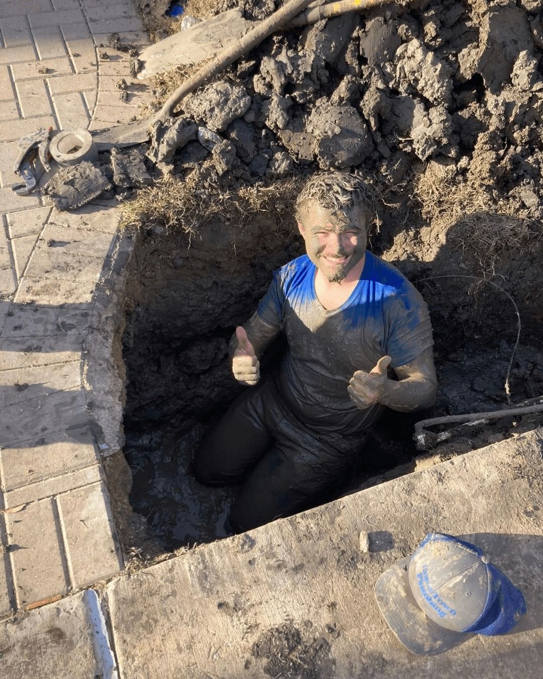 Man covered in mud gives thumbs up from a square hole beside a brick walkway.