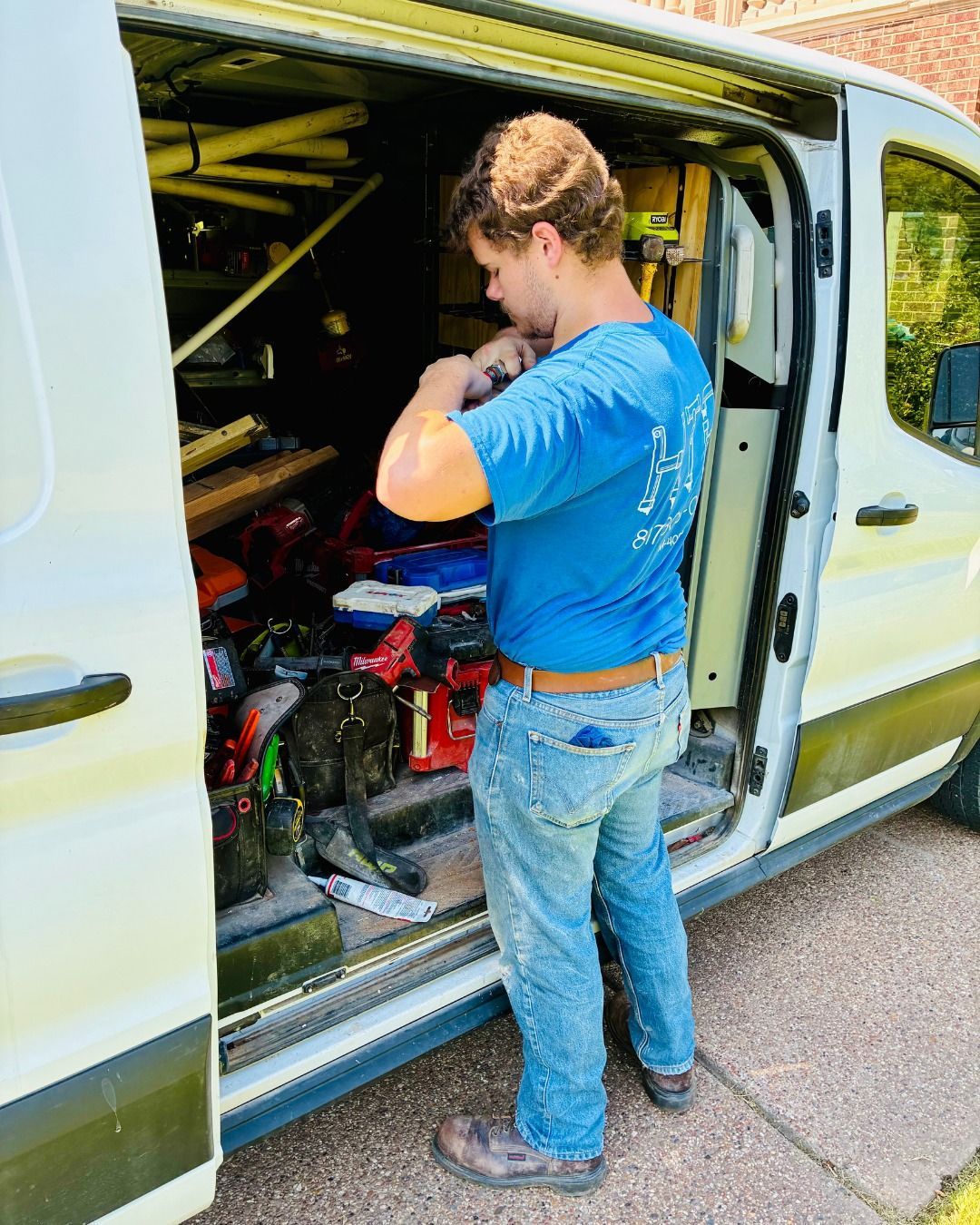 Person in a blue shirt and jeans stands by an open van, looking inside; tools are visible.