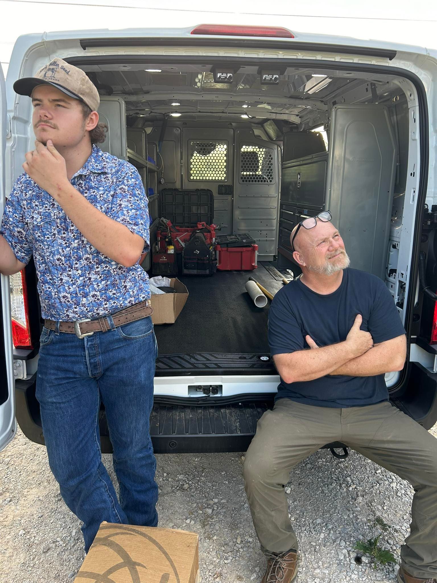 Two men near an open cargo van. One leans on the van, contemplating; the other sits, looking up.