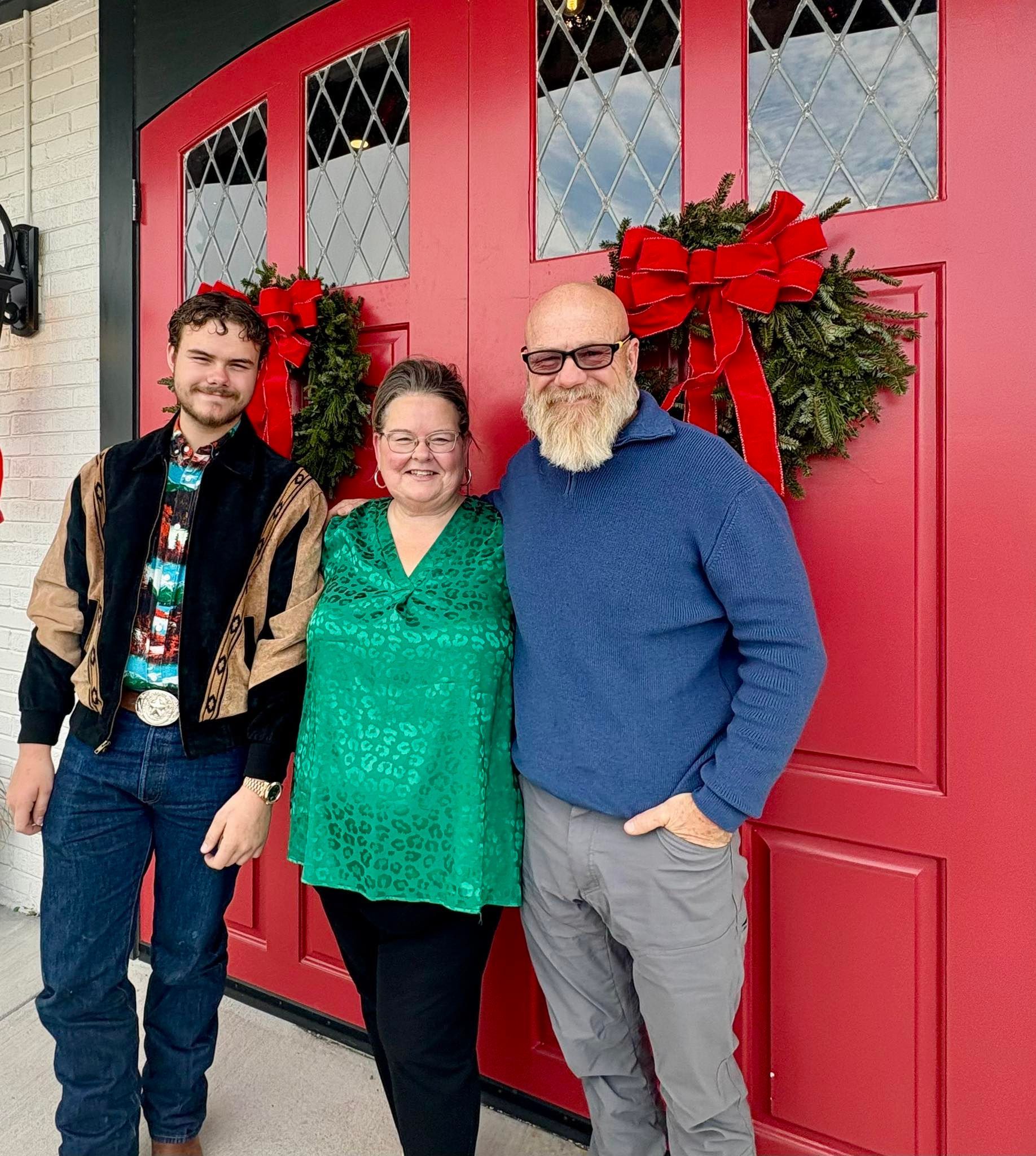 Three people pose in front of red doors with Christmas wreaths. The woman wears green, the man blue, the other a western jacket.