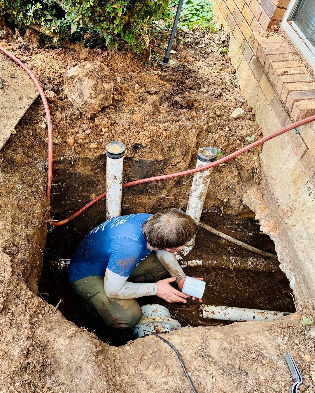Person examining pipes in an excavated area near a brick building.