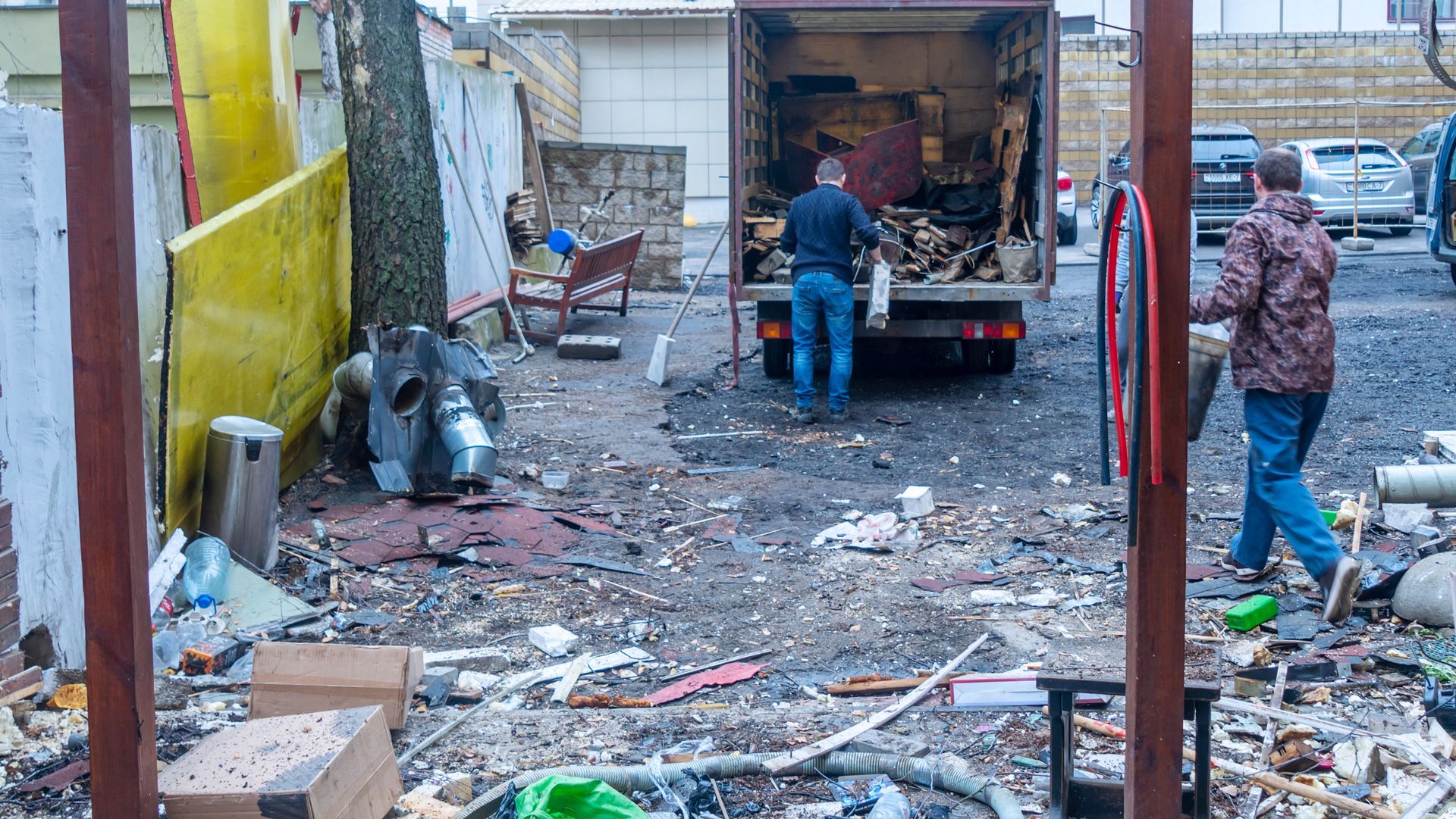 People loading a truck in a narrow, littered alley beside a yellow wall.