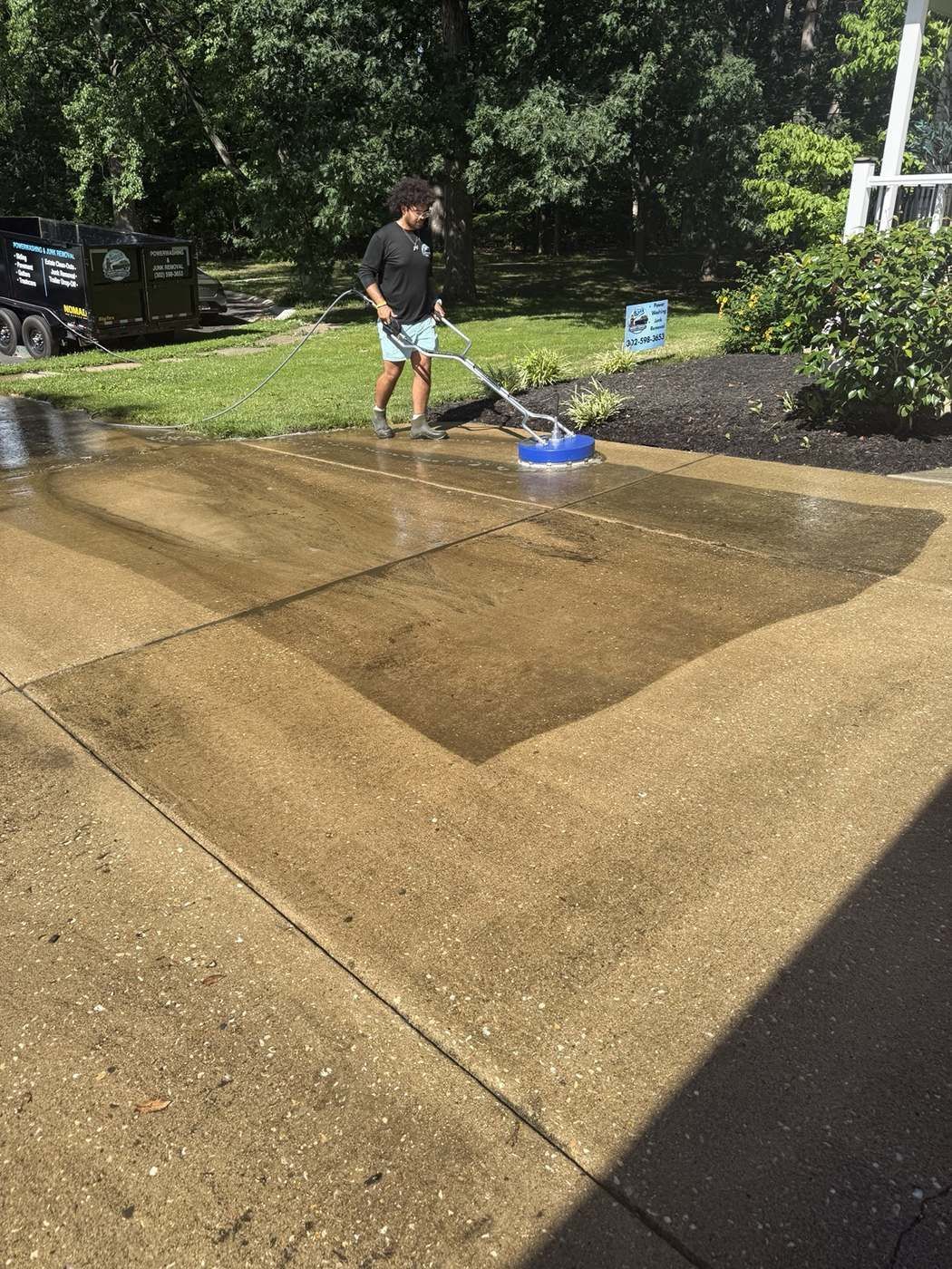 Technician surface cleaning a residential driveway with pressure washing equipment and branded trailer