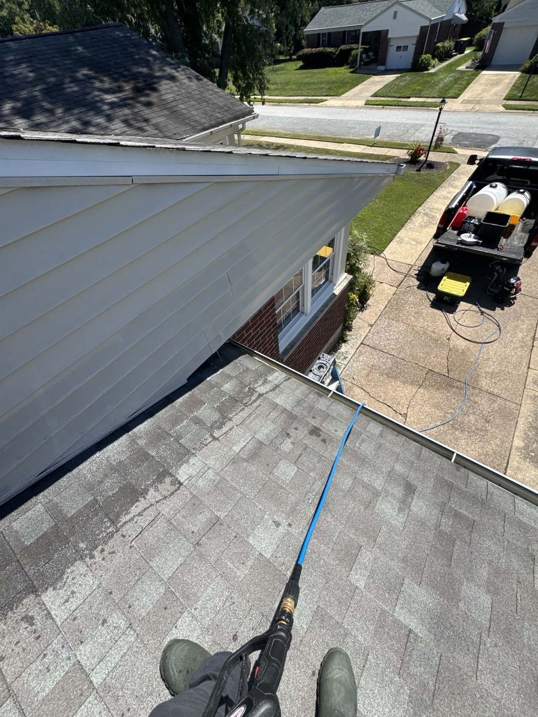 Technician on rooftop pressure washing gutters and siding with equipment truck below