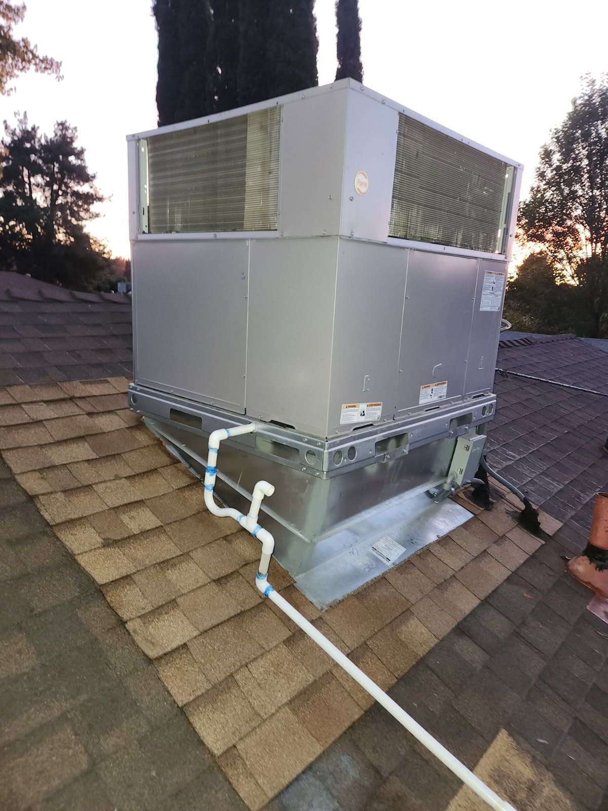 Rooftop HVAC unit on a shingled roof, with white PVC piping.