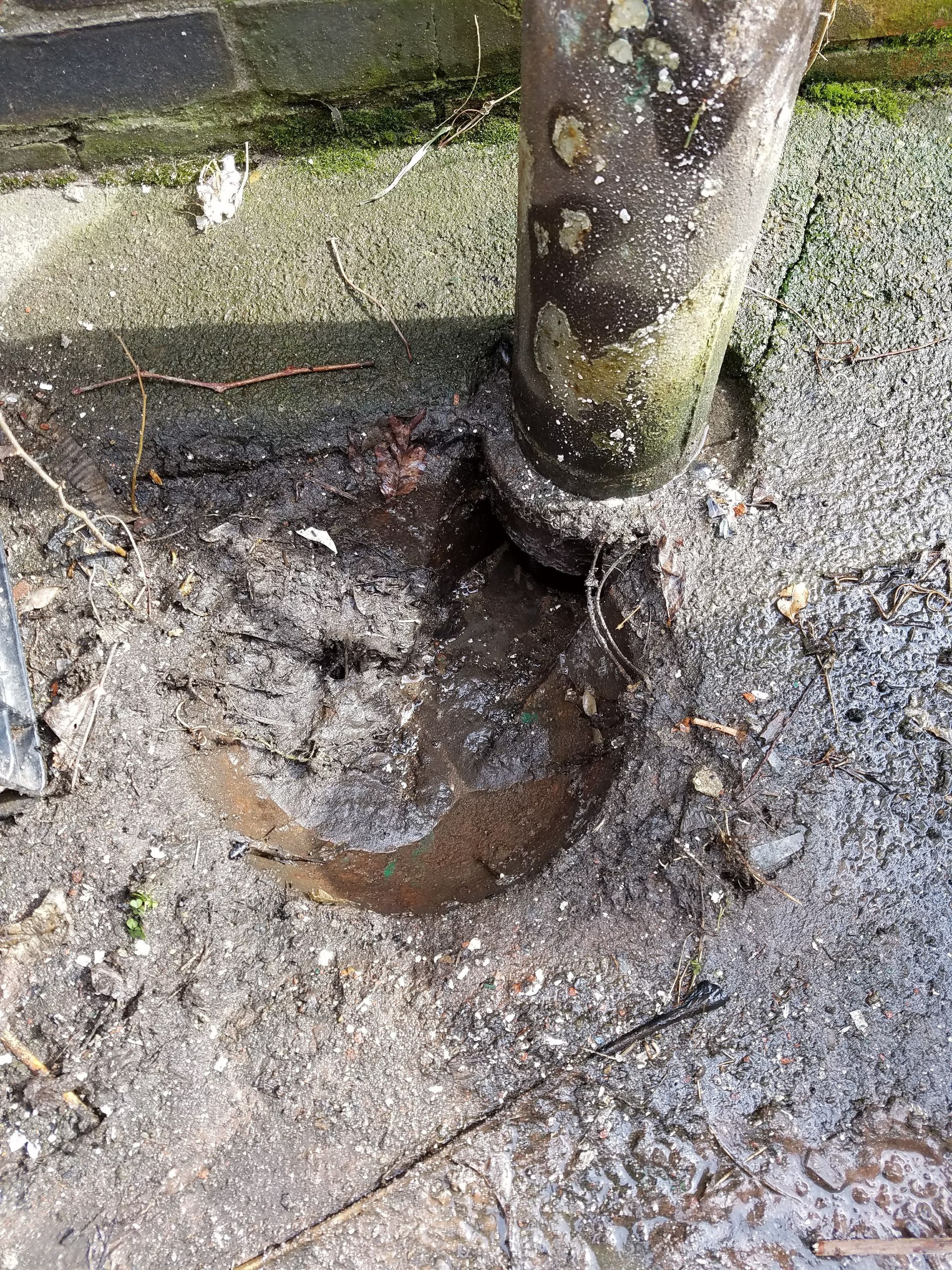 A rusty metal drainpipe connected to a broken semicircular metal section on dirty ground.