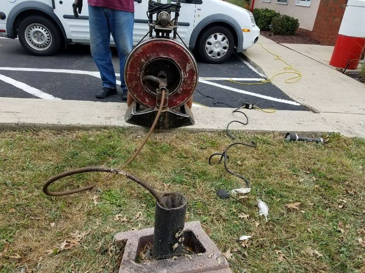 A man operates a sewer snake machine near a pipe in a parking lot.