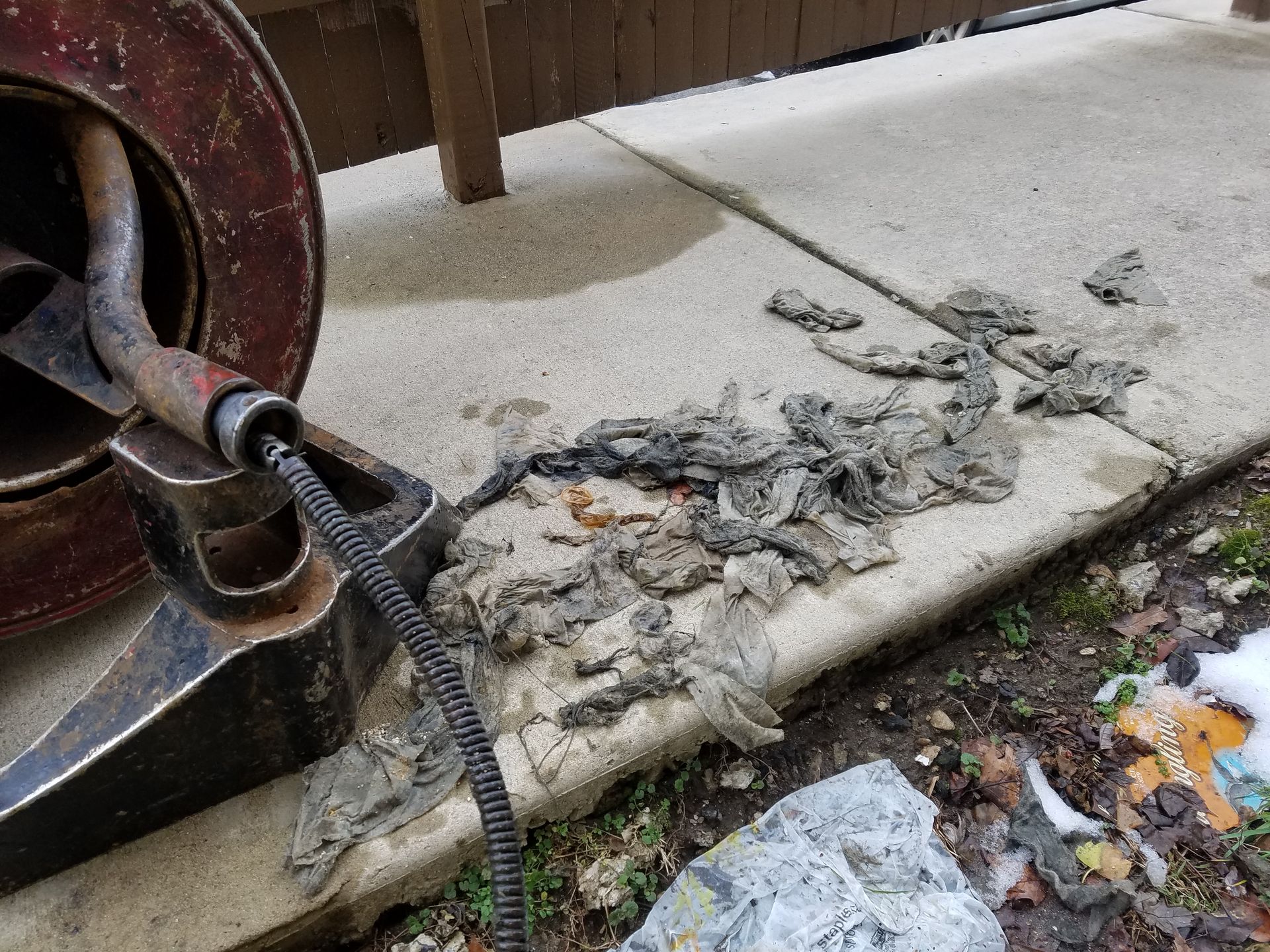 A drain snake beside a concrete walkway, extracting a mass of dark debris; outdoor setting.