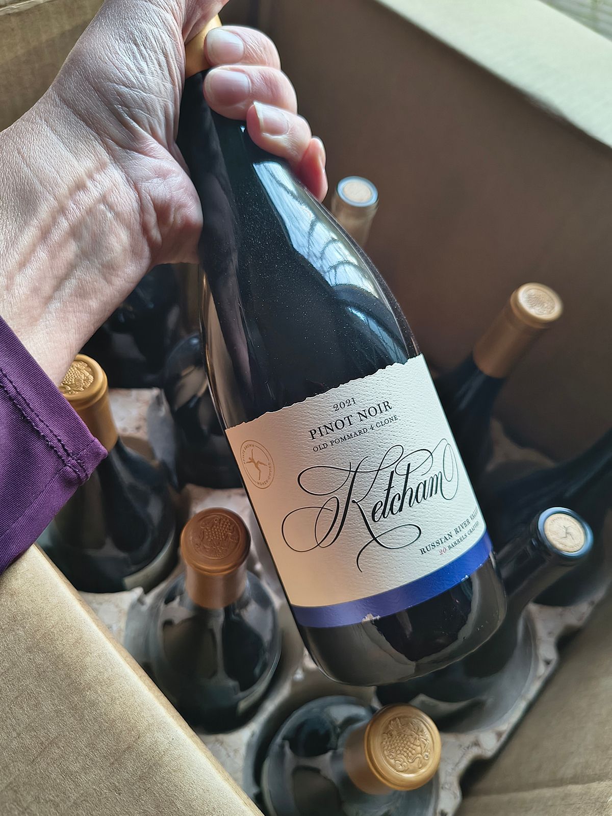 Hand holding a dark wine bottle in a cardboard box of assorted wine bottles