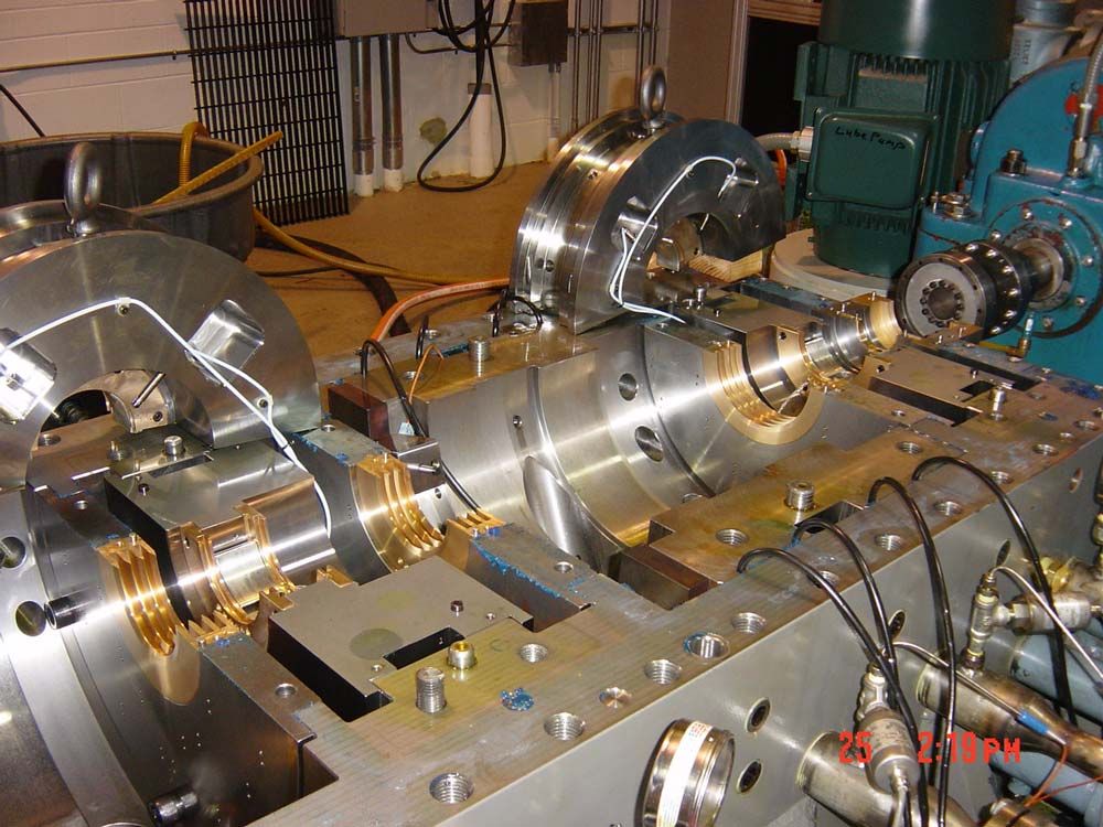 Polished metal machine assembly on a workbench, with gears, shafts, and wires in a workshop.