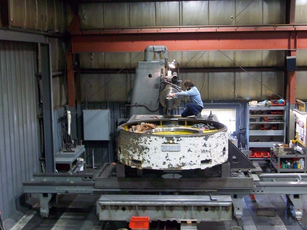 Worker repairing machinery in a workshop, leaning over a large metal machine on a platform.