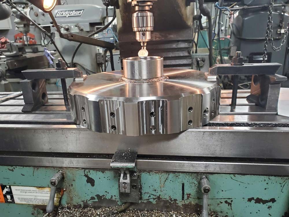 Polished metal machinery part being milled on an industrial workbench with a drill above it
