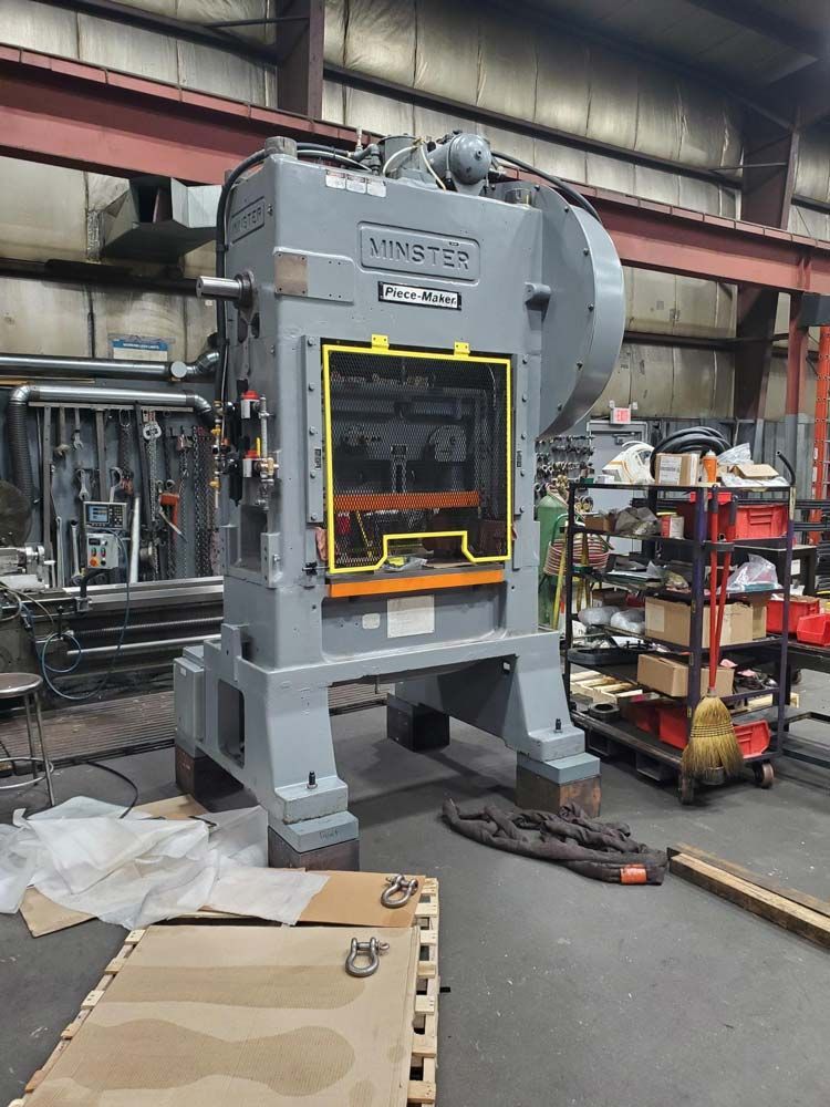 Large gray industrial press machine in a workshop, with yellow safety frame and shelving beside it.