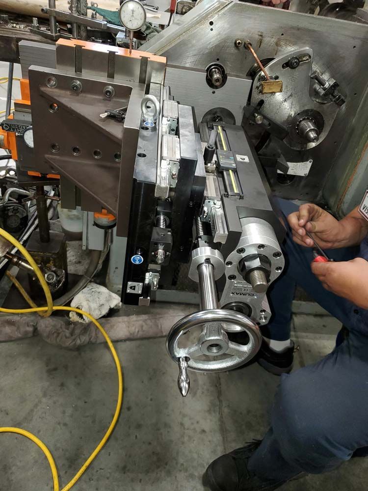 Worker operating a metal lathe in a workshop, with tools and cables around the machine.