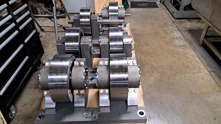Rows of shiny metal cylindrical parts mounted on a factory floor pallet, with shelves in the background